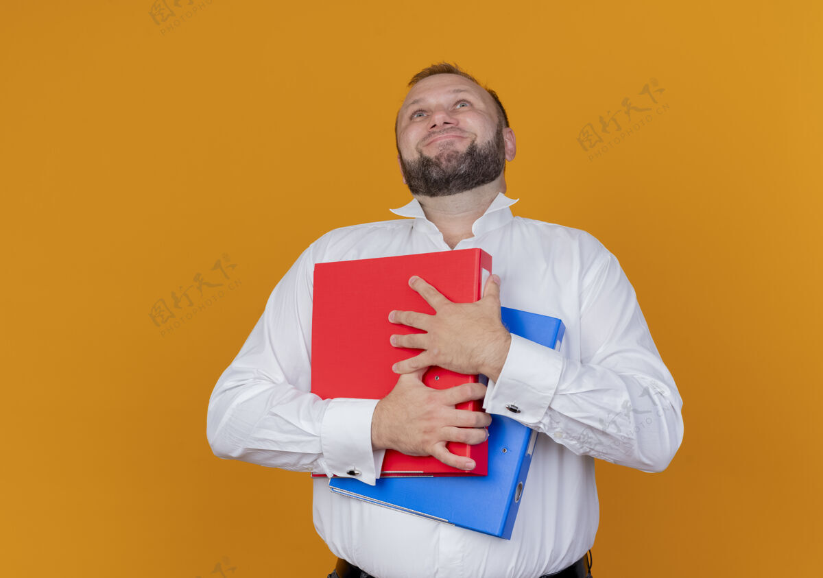 男人一个留着胡子的男人穿着白衬衫 举着文件夹 站在橘色的墙上 脸上挂着幸福的表情抱穿脸