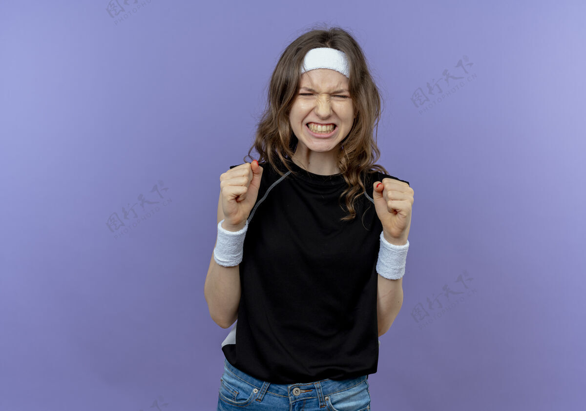 野性身穿黑色运动服的年轻健身女孩 头上戴着头巾 站在蓝色的墙壁上疯狂地握紧拳头握紧健身拳头