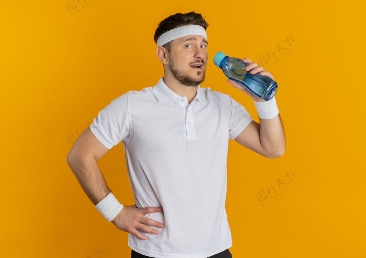 微笑身穿白衬衫 头箍着水瓶 脸上带着微笑的年轻健身男子站在橙色背景下看着摄像机拿着瓶子站着