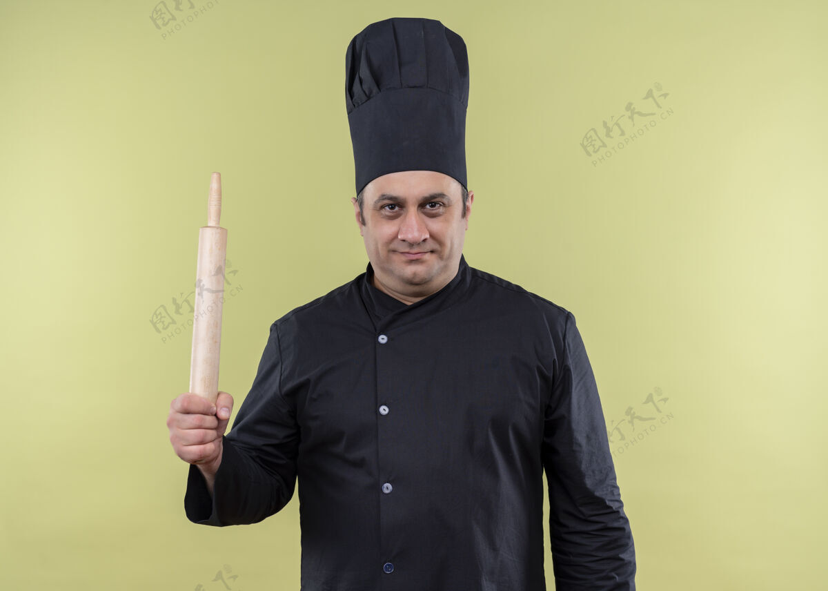 男性男厨师身穿黑色制服 头戴厨师帽 手持擀面杖 站在绿色背景下 自信地看着镜头黑色厨师帽子