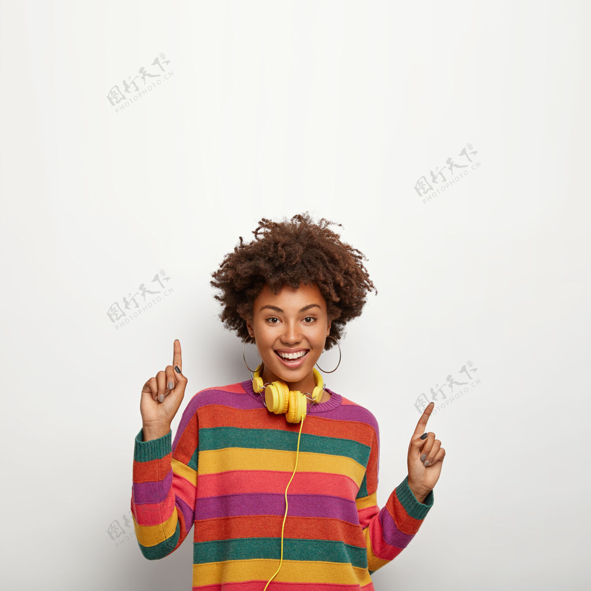 多彩美丽无忧无虑的时髦女孩 非洲式发型 靠着白墙 向上指 在这里说你的文字 用黄色耳机听喜欢的音乐 穿着条纹彩色套头衫休闲欢乐欢乐