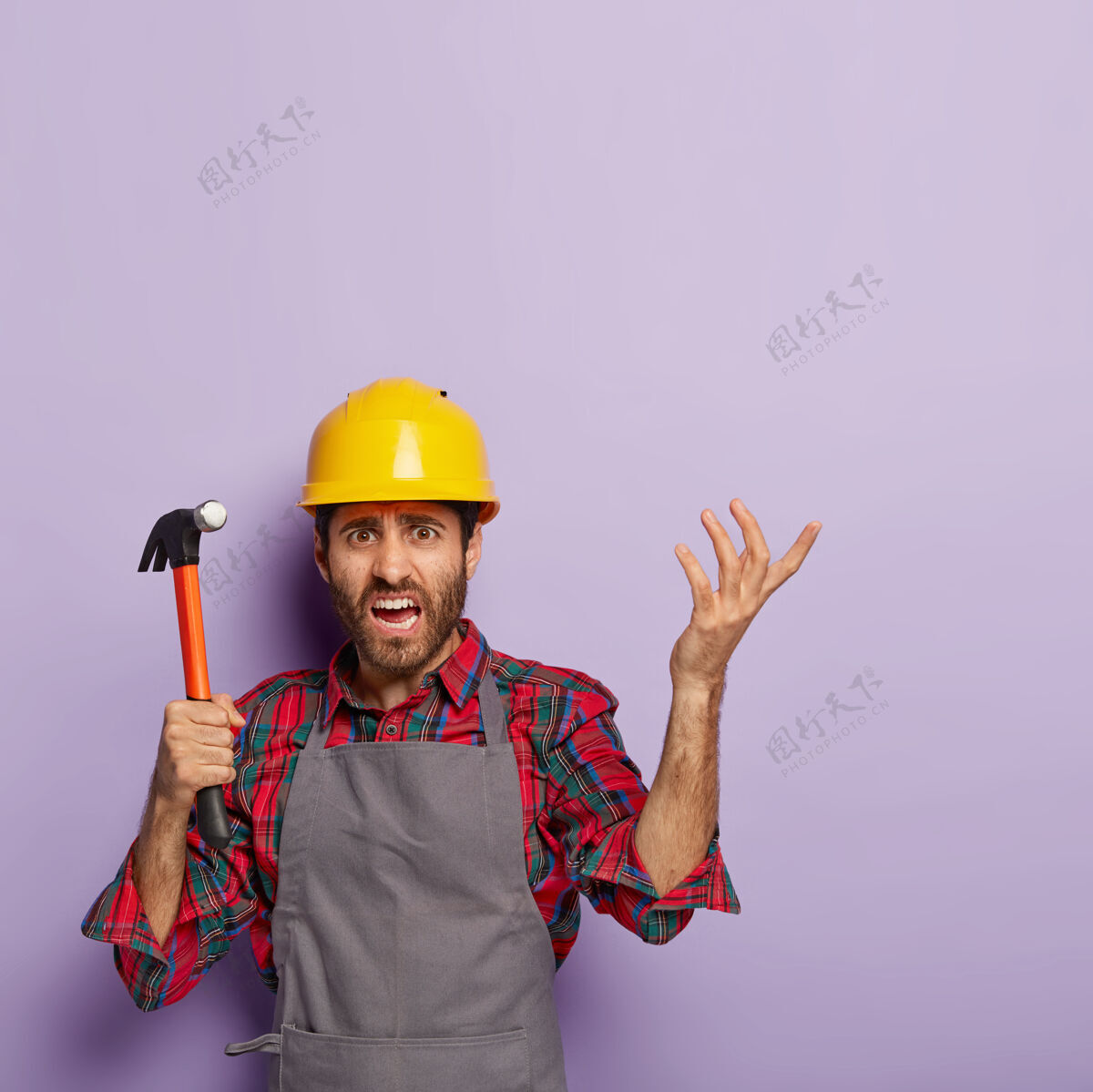 忙碌失意修理工的垂直镜头有着不解的表情 手里拿着锤子 义愤填膺地举起手臂 穿着工装 不懂修理什么工具建筑工人不快乐