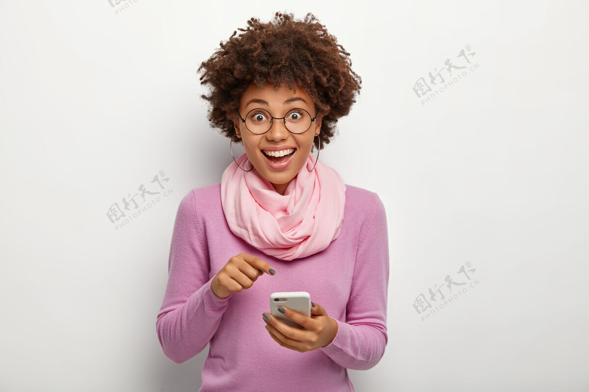 指秀发清爽 面容姣好的女性 指着手机 戴上新的现代应用 表情愉快 戴着矫正视力的眼镜 紫色套头衫和丝巾站应用程序使用
