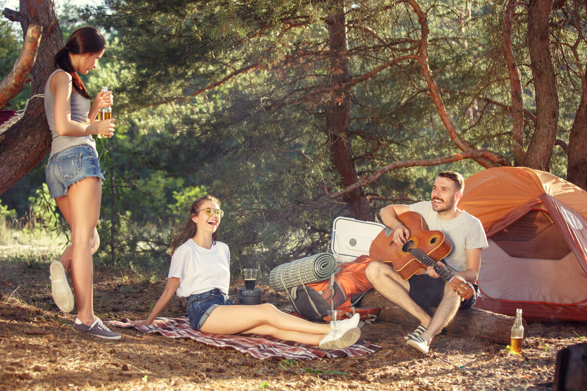 一起聚会 男女露营在森林里他们放松 唱着歌对抗绿草度假 夏天 冒险 生活方式 野餐的概念白天食物篝火