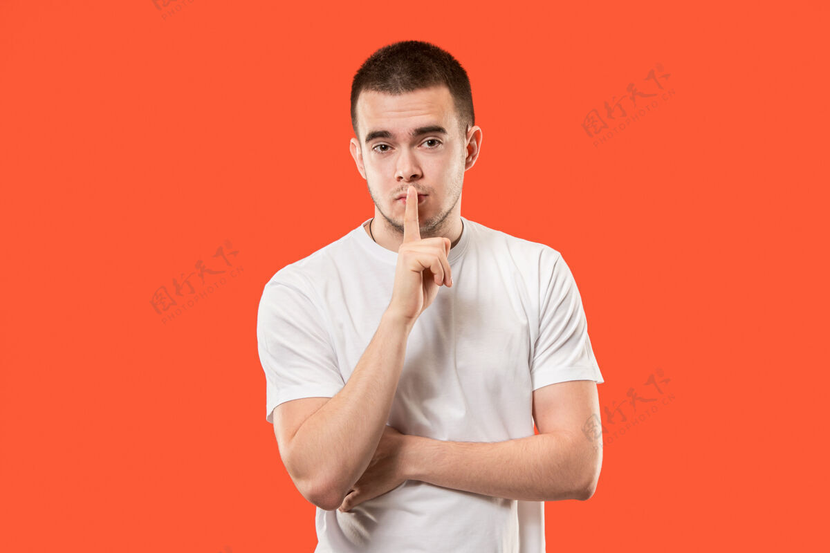 秘密那个年轻人在橙色的背景上用手在背后低语着一个秘密表情成人手势