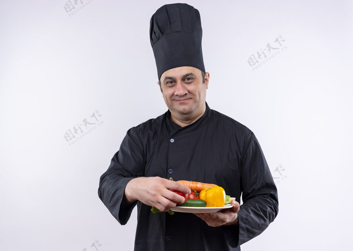 男男厨师身穿黑色制服 头戴厨师帽 手持盛有新鲜蔬菜的盘子 站在白色背景下 面带微笑地看着镜头盘子穿看