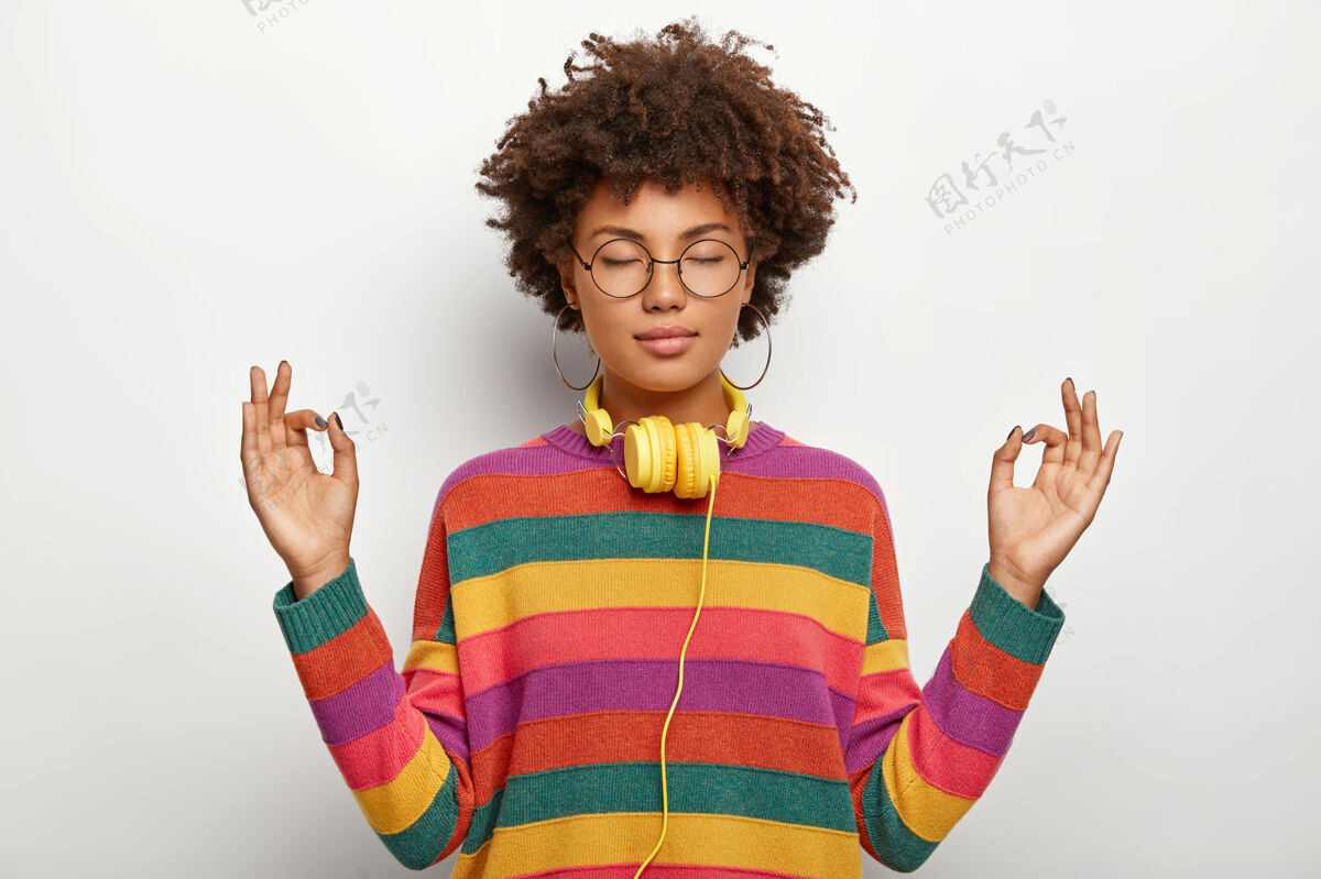 室内非洲发型的放松美女在室内冥想 闭上眼睛 戴着耳机听悦耳的音乐 戴着眼镜 耳环 条纹套头衫肢体语言女性放松平衡