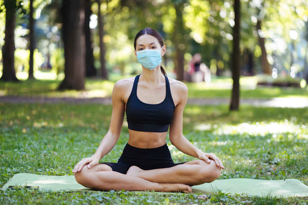 健康戴着医用防护面具的运动型年轻女子 早上在公园里做瑜伽 在瑜伽垫上进行女子训练防护运动瑜伽