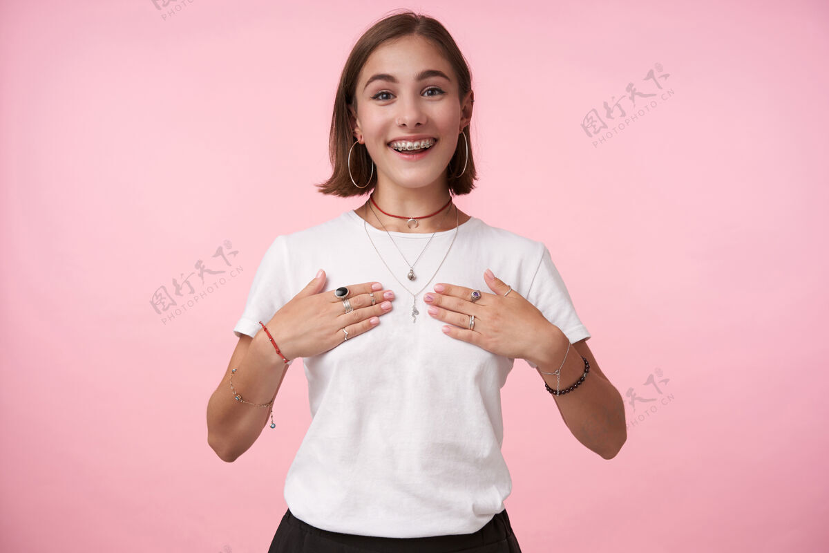 手掌摄影棚拍摄的年轻可爱的深色头发的女性鲍勃剪头发愉快地看着前面 同时保持她的胸部手掌 隔离在粉红色的墙壁积极姿势休闲