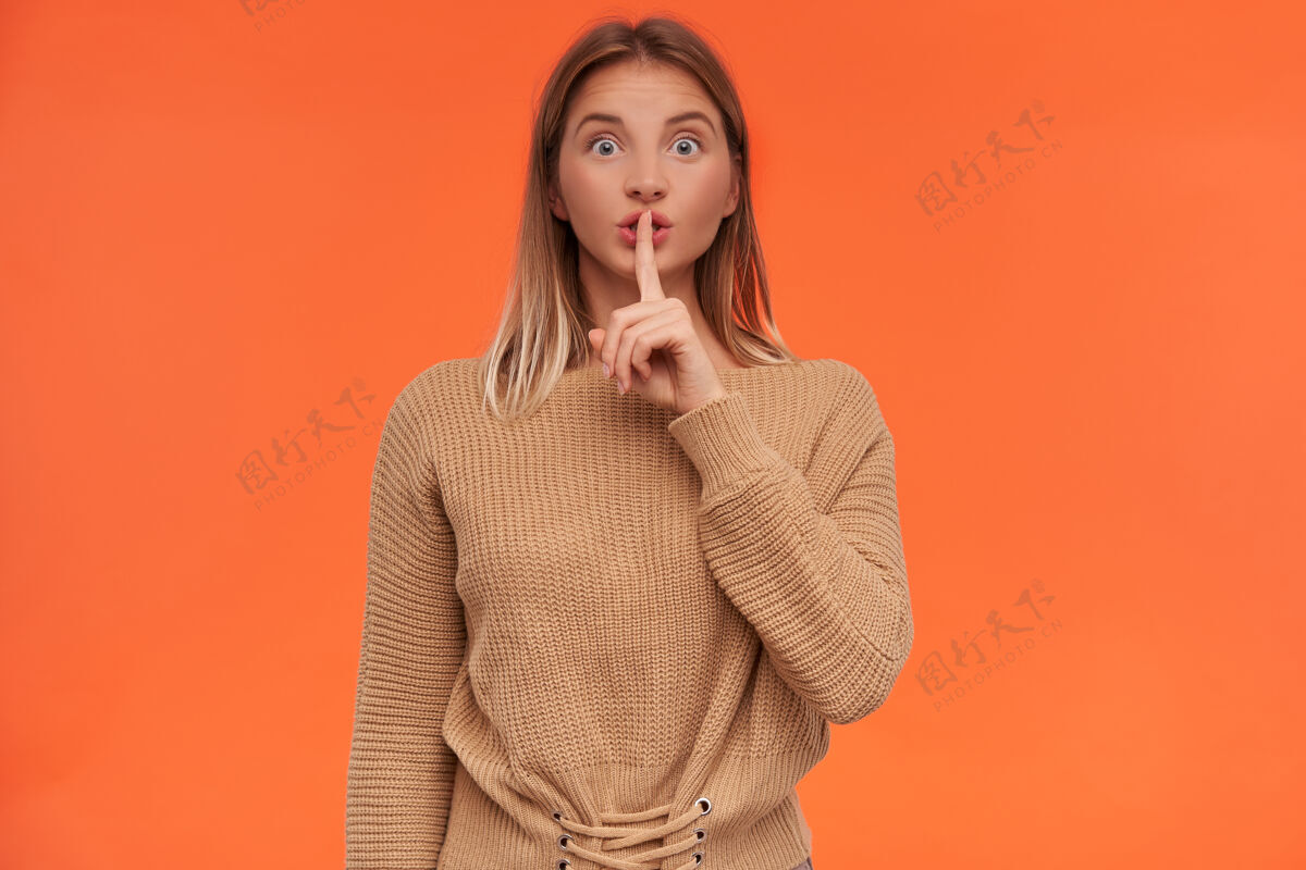 表情兴奋的年轻蓝眼睛白头发的女人举起手在安静的手势 她的嘴 同时看着前面的情绪 站在橙色的墙毛衣衣服肖像