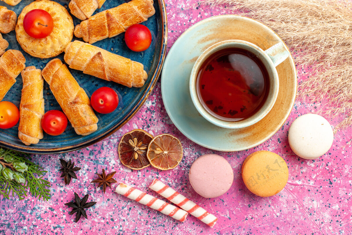 甜点顶视图甜甜可口的百吉饼在托盘内与新鲜酸李子法国马卡龙和一杯茶在淡粉色的桌子上派杯子早餐