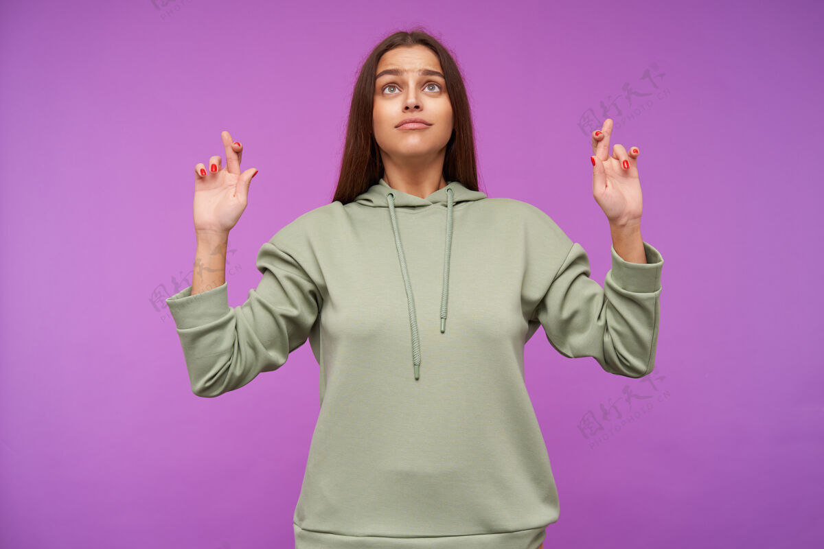 运动站在紫色的墙上 仰望着年轻的碧眼黑发女人 用手指交叉着自然的妆容 祈求好运宽松女士交叉