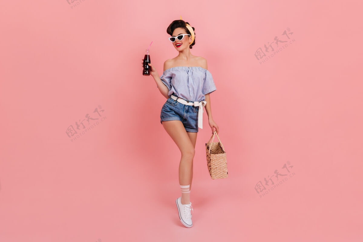 女士身材匀称的小姑娘手里拿着一瓶苏打水 面带微笑迷人的欧洲女人拿着一个夏天的袋子 在粉色的背景上摆姿势优雅玩苏打水