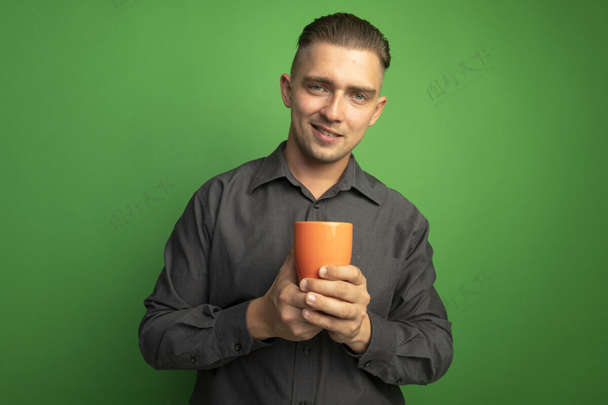 人身穿灰色衬衫的年轻帅哥拿着橘色的马克杯望着前面 微笑着自信地站在绿色的墙上英俊人微笑