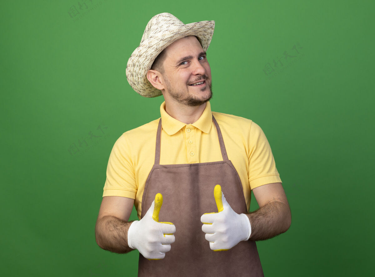 人年轻的园丁穿着连体衣 戴着帽子 戴着工作手套 站在绿色的墙上 面带微笑地竖起大拇指 看着前面人市民连身衣
