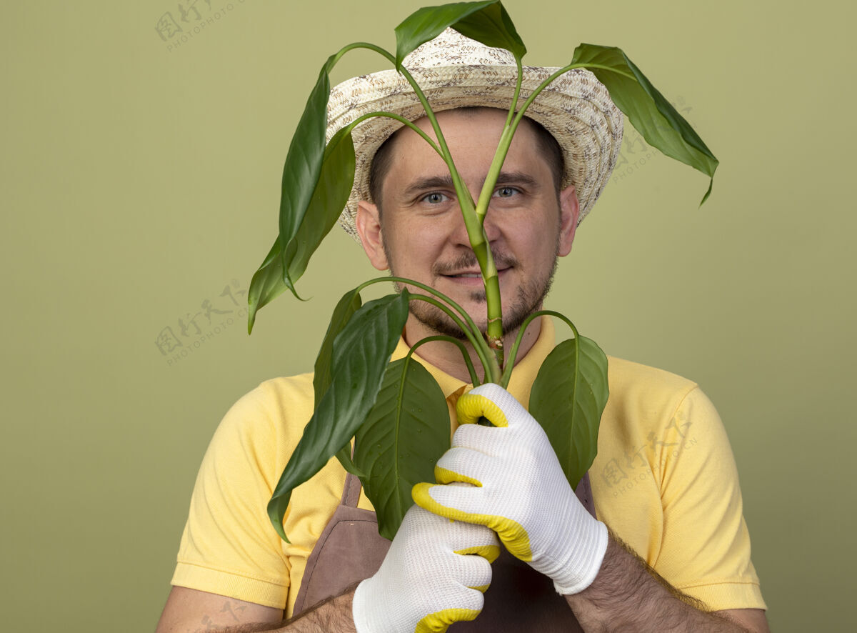 微笑年轻的园丁 穿着连体衣 戴着帽子 戴着工作手套 手里拿着一株植物 站在明亮的墙上 面带微笑地看着前方抱连身衣人