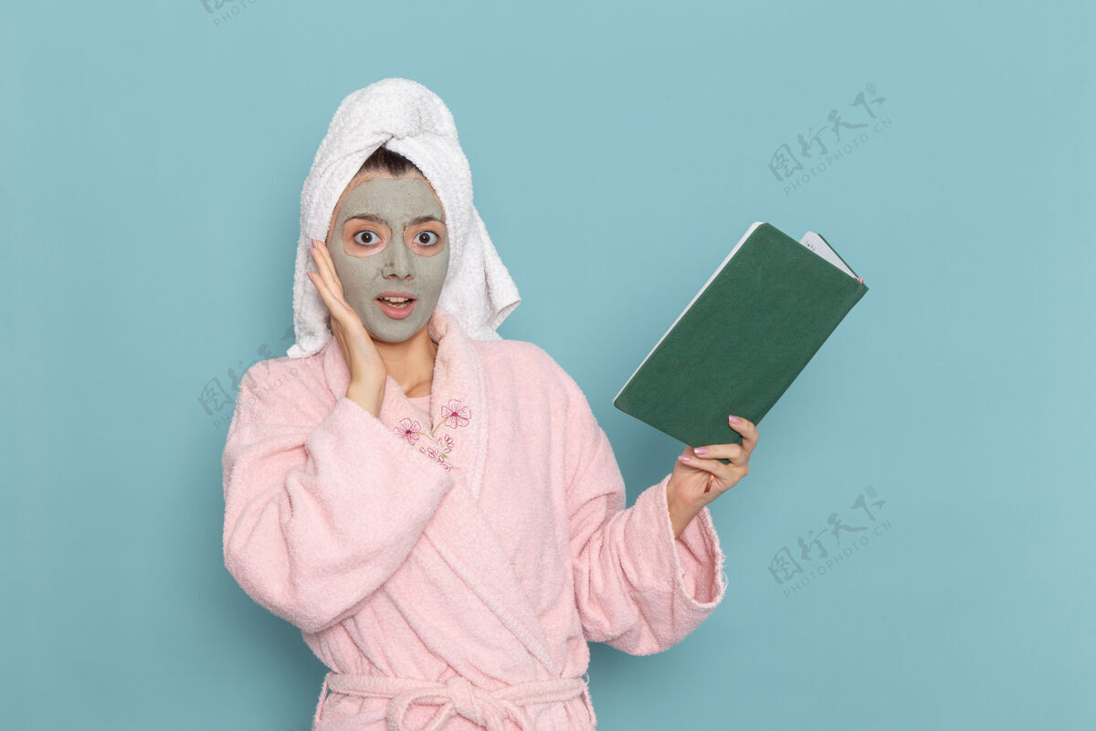 淋浴正面图身着粉色浴袍的年轻女性手持抄写本在蓝色墙壁上阅读美丽的水浴霜自理淋浴查看人浴袍