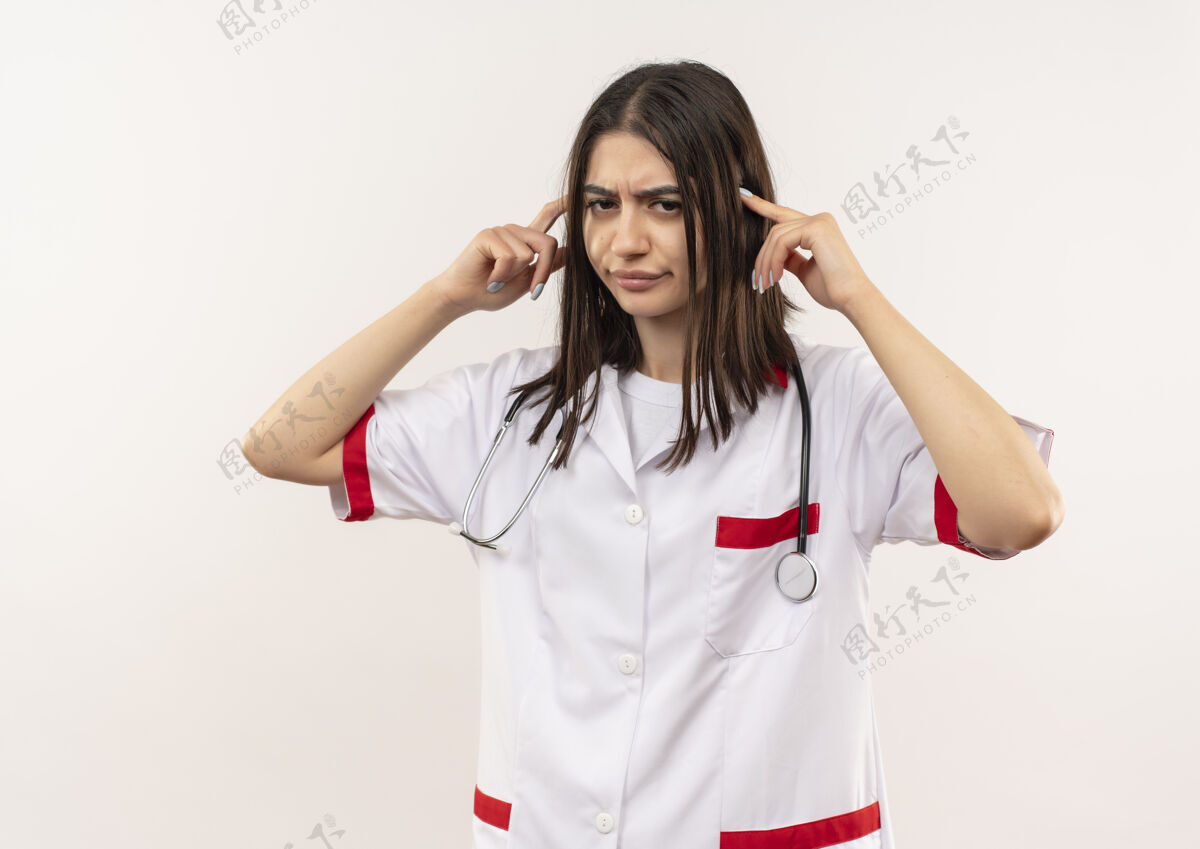 女人年轻的女医生穿着白大褂 脖子上戴着听诊器指着太阳穴 站在白墙上显得又累又无聊太阳穴姿势无聊