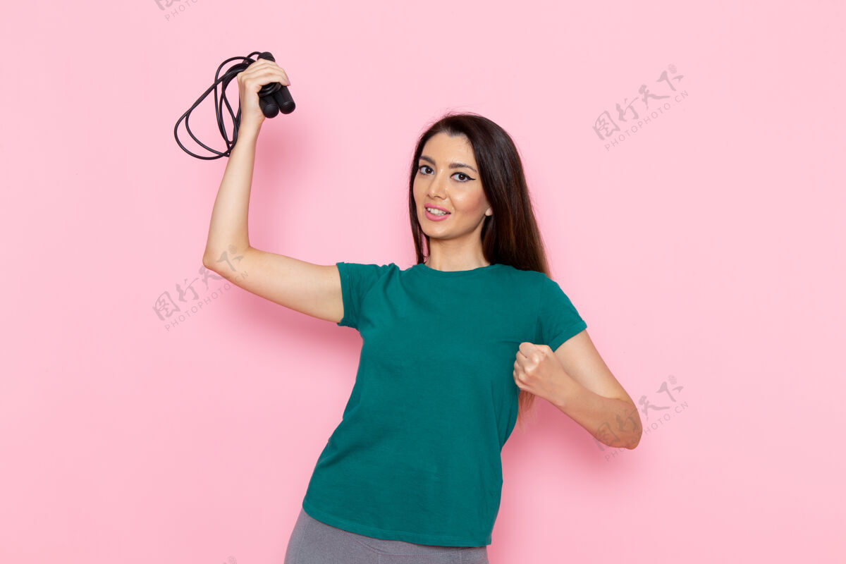 浅粉色正面图身着绿色t恤的年轻女性手持浅粉色墙壁上的跳绳腰部运动锻炼美丽苗条女性运动腰部查看锻炼