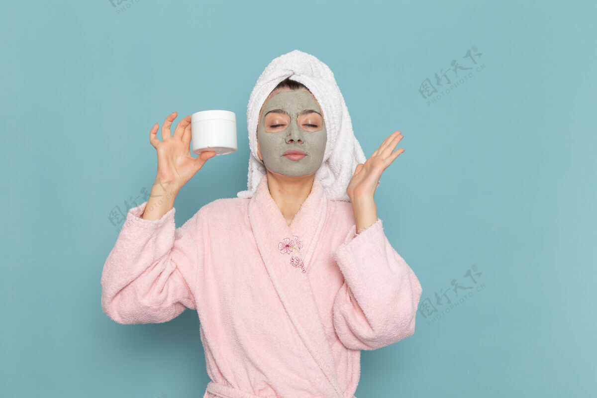 奶油正面图身着粉色浴袍的年轻女性手持面霜在蓝色墙壁上淋浴清洗美容自护面霜自理人举行
