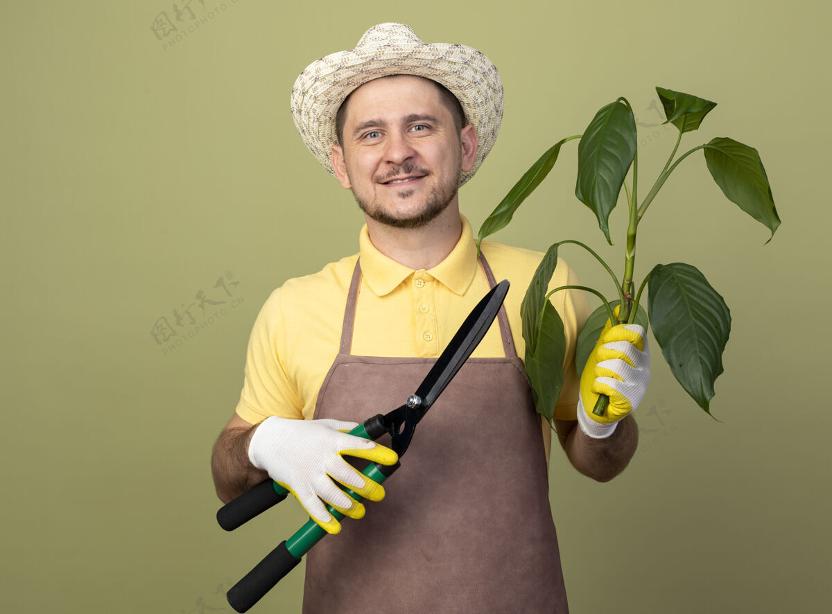 制服年轻的园丁身穿连体衣 戴着帽子 戴着工作手套 手里拿着植物和树篱修剪器 面带微笑地站在明亮的墙上看着前方连身衣植物脸
