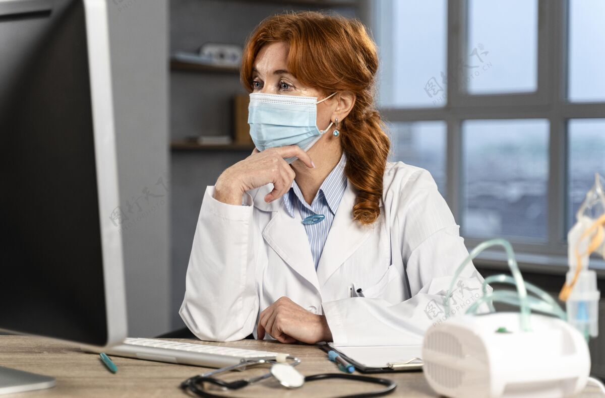 女人女医生戴着医用面罩坐在办公桌前看电脑面罩职业临床医生