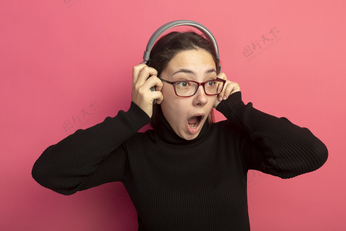 边一个穿着黑色高领毛衣戴着眼镜戴着耳机的年轻漂亮女人站在粉红色的墙上惊讶地张大了嘴张开耳机黑色