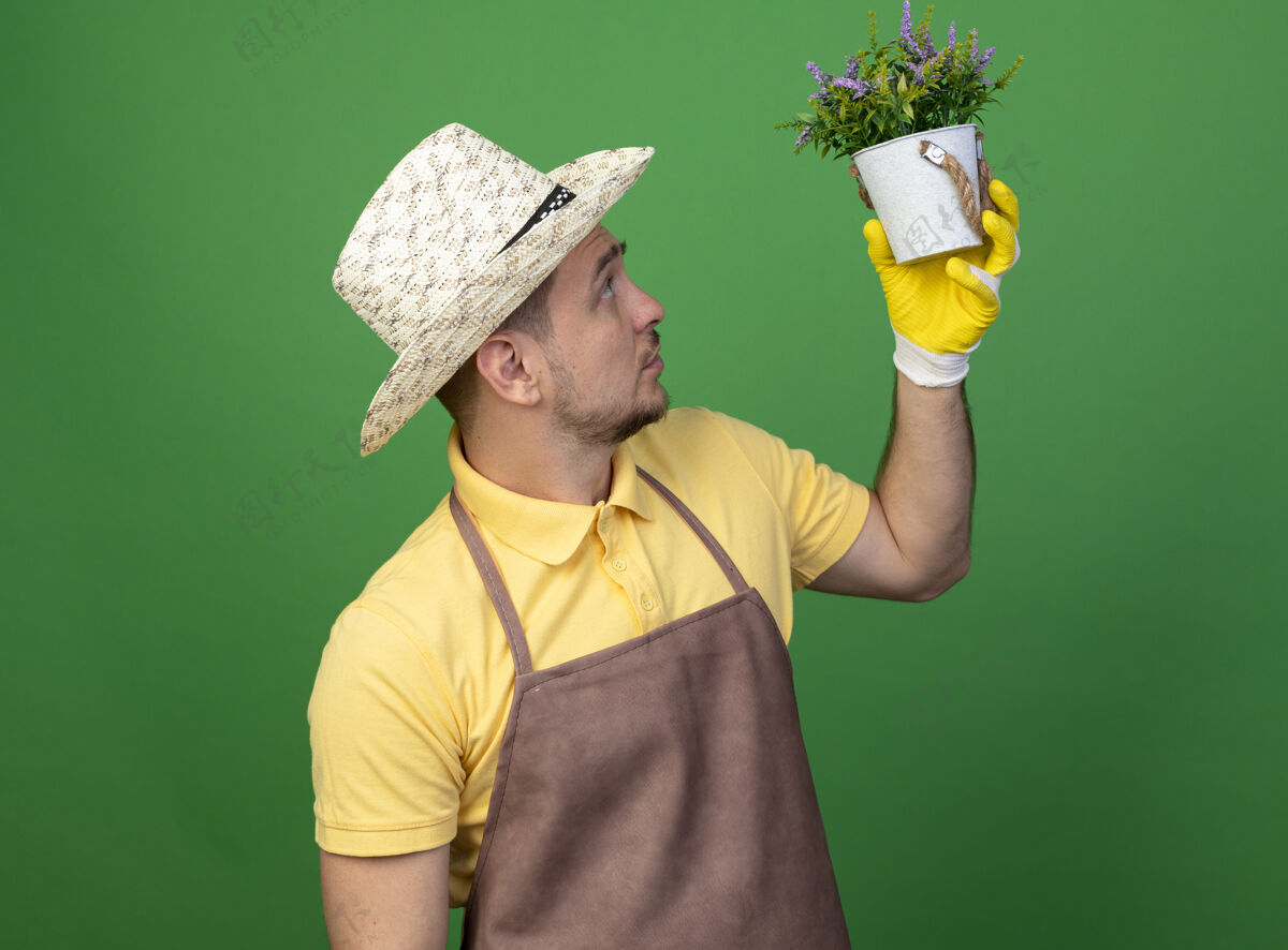 帽子年轻的园丁穿着连体衣 戴着帽子 戴着工作手套 手里拿着盆栽植物 站在绿色的墙上好奇地看着它穿制服人