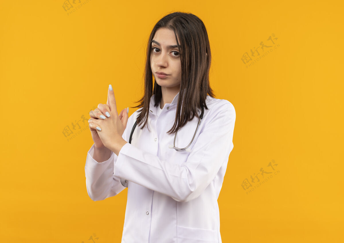 外套年轻的女医生穿着白大褂 脖子上戴着听诊器 用手指朝前做着手枪或枪的手势 严肃的脸站在橙色的墙上医生医院年轻人