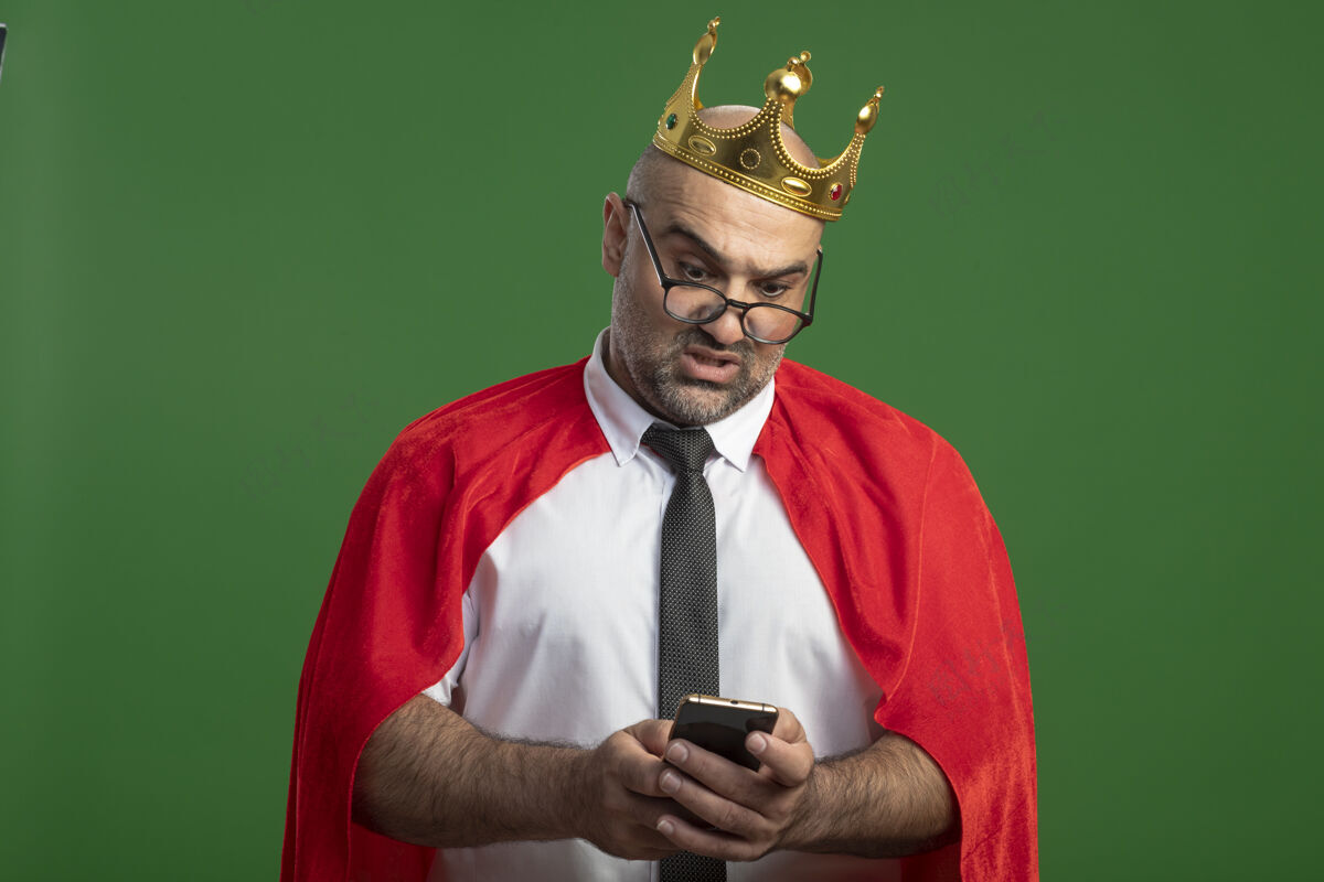 人穿着红色斗篷戴着皇冠的超级英雄商人站在绿色的墙上 用智能手机看着困惑商人手机面部表情