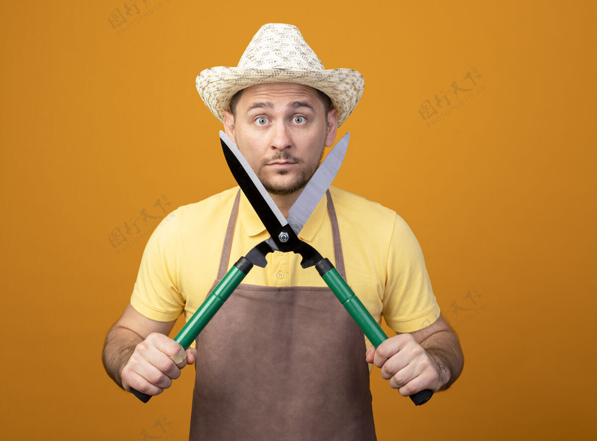 工人年轻的园丁穿着连体衣 戴着帽子 手持树篱修剪器 站在橘色的墙上 看着前方 感到惊讶和惊奇剪子惊人花园