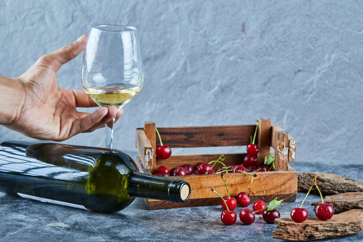 手一个女人拿着一杯白葡萄酒和一盒樱桃放在蓝色的表面上饮料有机木头