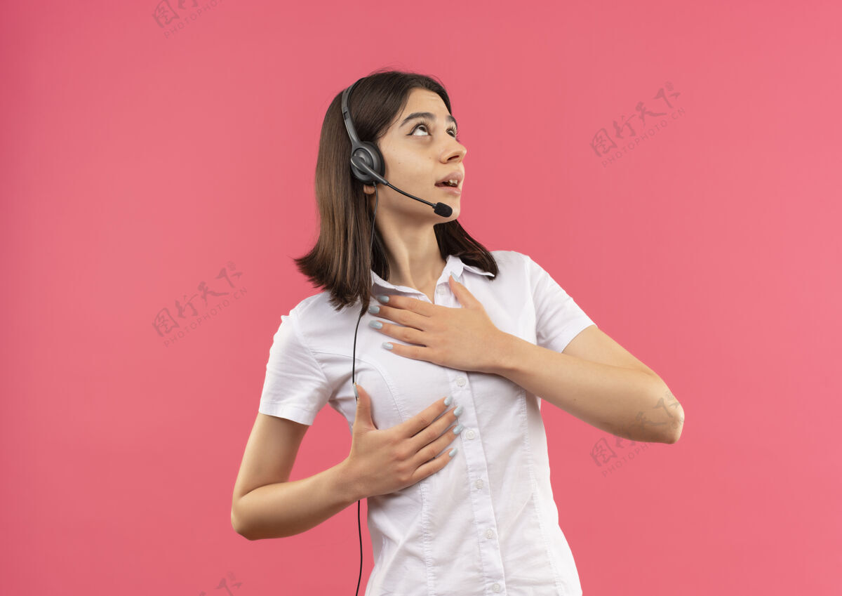 手势一个穿着白衬衫戴着耳机的年轻女孩 手放在胸前 站在粉红色的墙上焦急地看着一边旁听女性担心