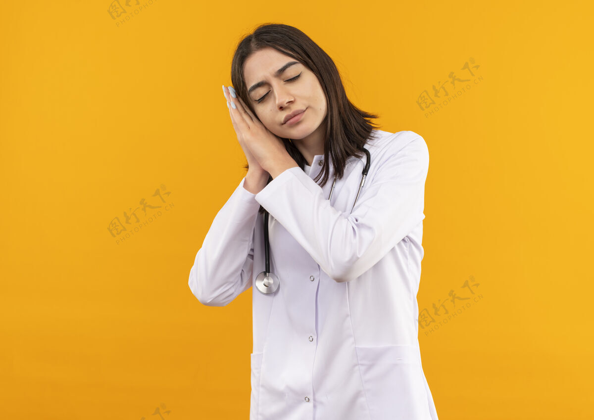公民年轻的女医生穿着白大褂 脖子上戴着听诊器 双手合十 头靠在手心上 站在橘色的墙上想睡觉 看起来很累疲倦女人年轻