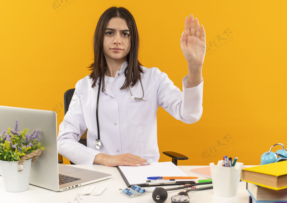 停止年轻的女医生穿着白大褂 手持听诊器 面带严肃的表情 手拿着笔记本电脑和文件坐在桌子旁 站在橙色的墙上 朝前面看坐着笔记本电脑外套