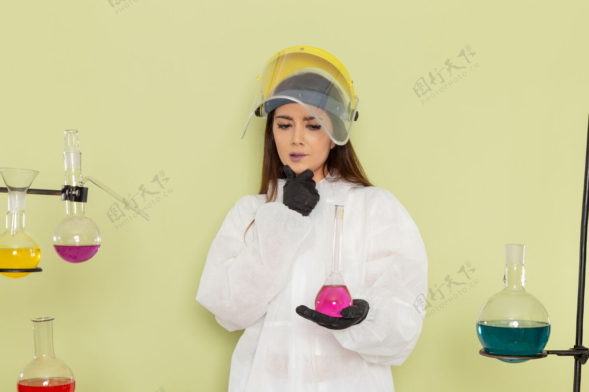 持有正面图身着特殊防护服的年轻女化学家手持粉色溶液 思考绿色墙化学工作女科学实验室科学服装特殊