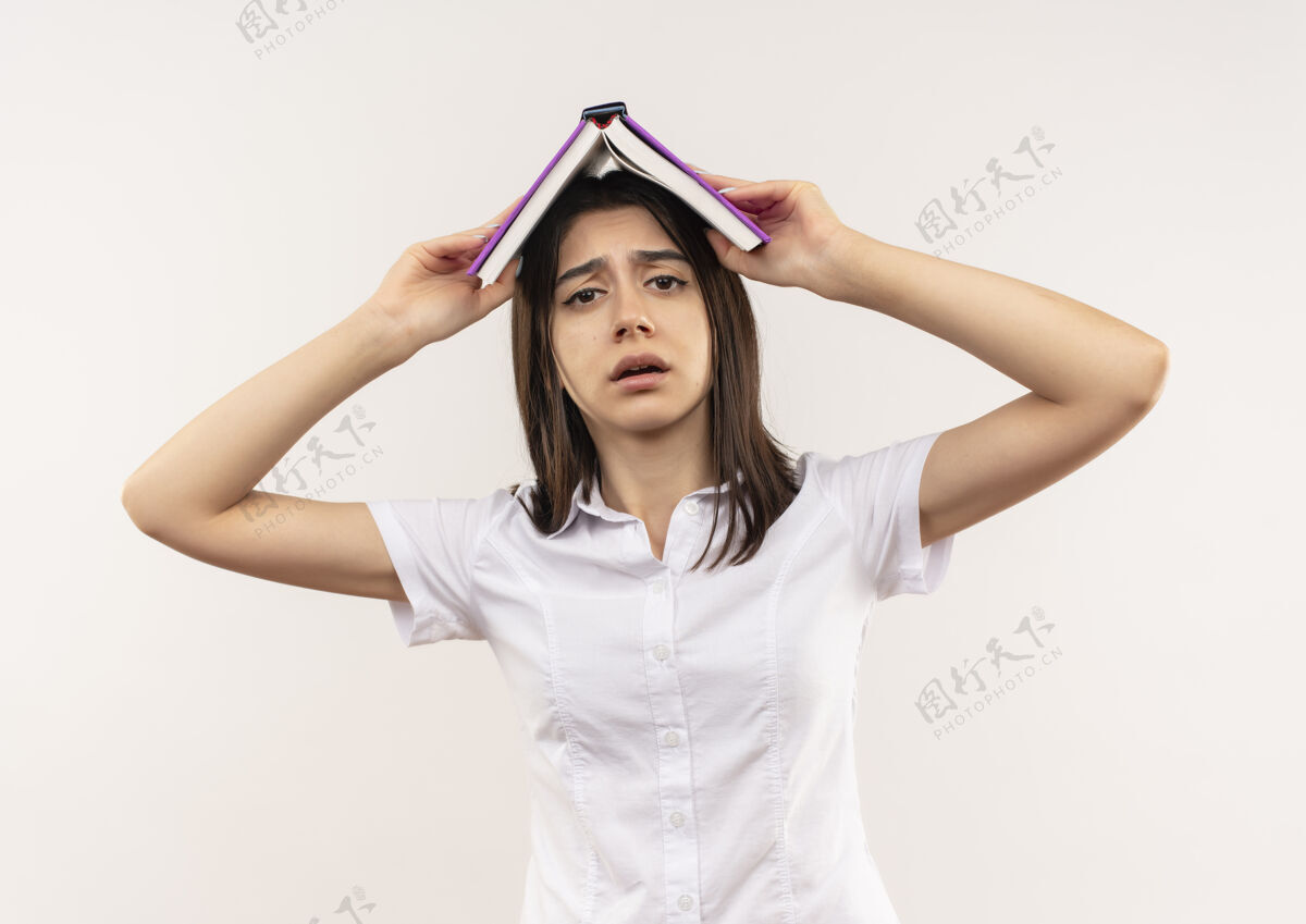 女穿着白衬衫的年轻女孩头上举着一本打开的书 站在白色的墙上 看上去很困惑衬衫站着开