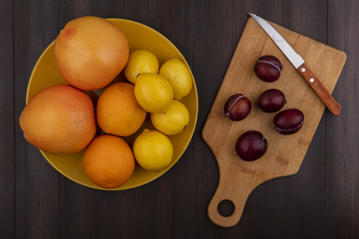 食物用刀在砧板上俯瞰李子 在木制背景上的碗里放着柠檬 橘子和葡萄柚甜食碗板