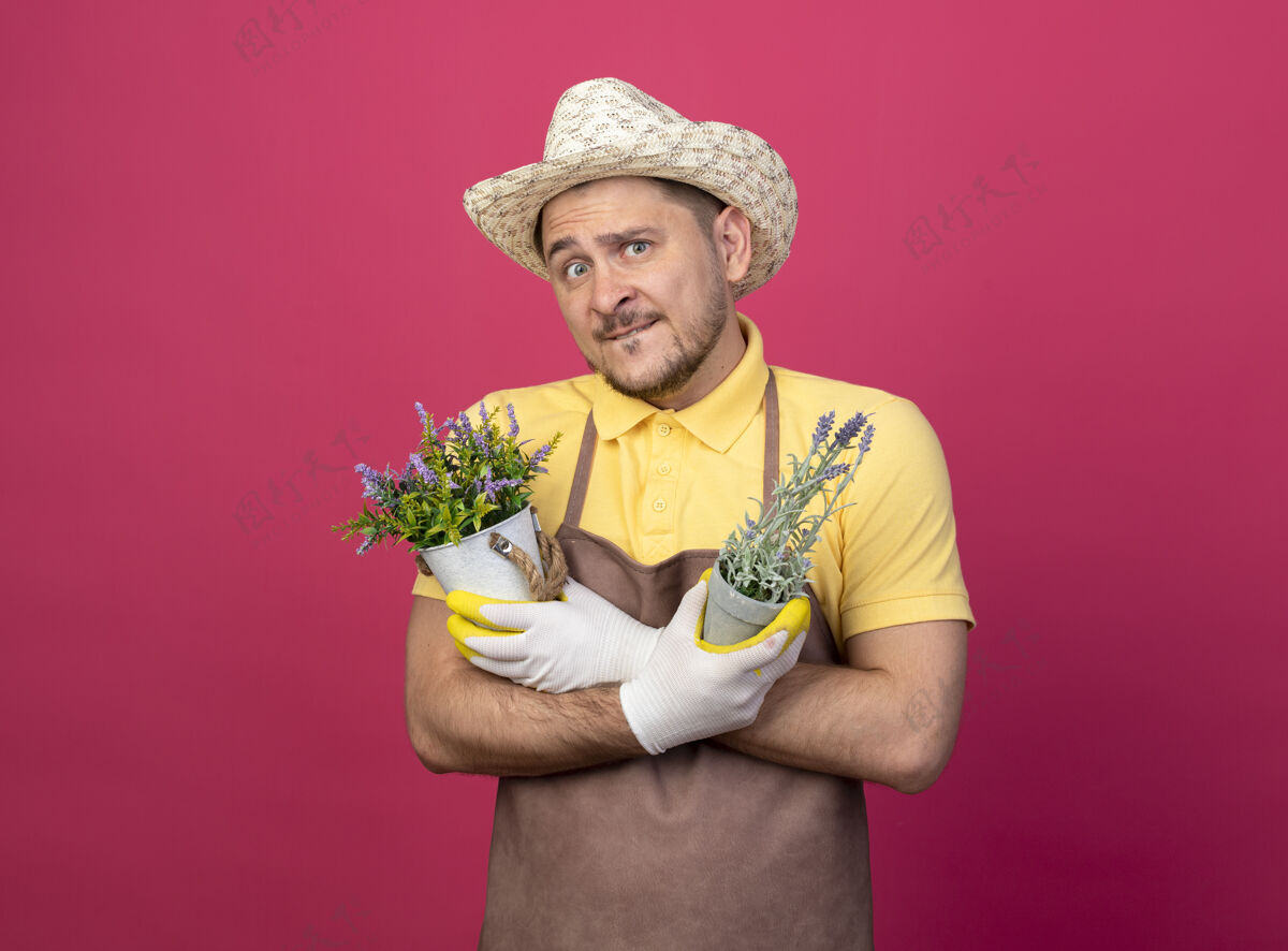 植物年轻的园丁穿着连体衣 戴着帽子 戴着工作手套 手里拿着盆栽植物 站在粉色的墙上困惑地看着前面人手套拿着