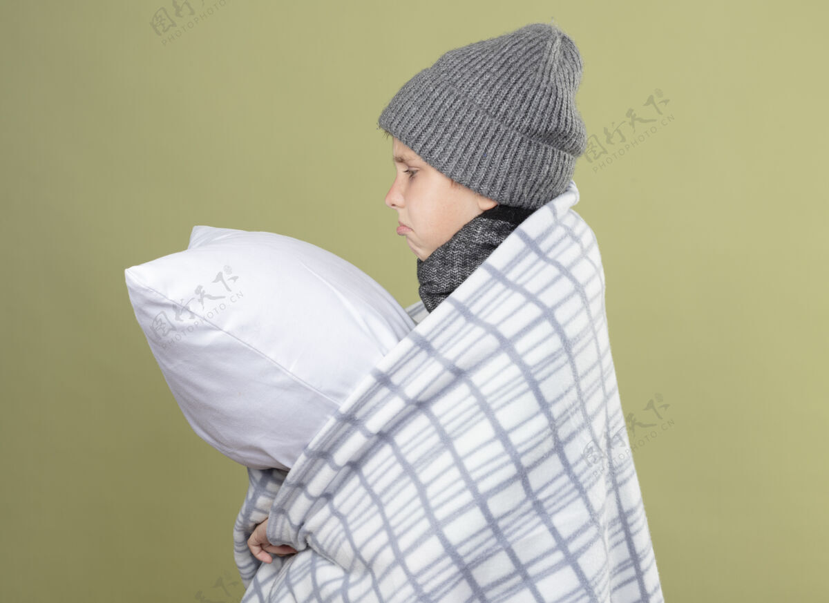 帽子生病的小男孩 戴着暖和的帽子 裹着毯子 抱着枕头 身体不舒服 站在一边 不开心 病倒在轻墙上小毯子感觉