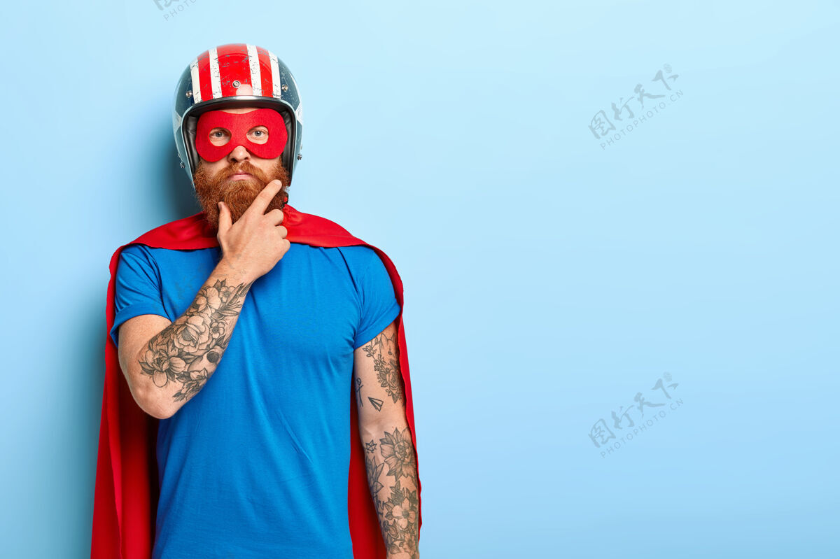 胡须人和超级权力概念严肃的男人 红色浓密的胡须 戴着头盔和红色的超级英雄斗篷力量男性面具
