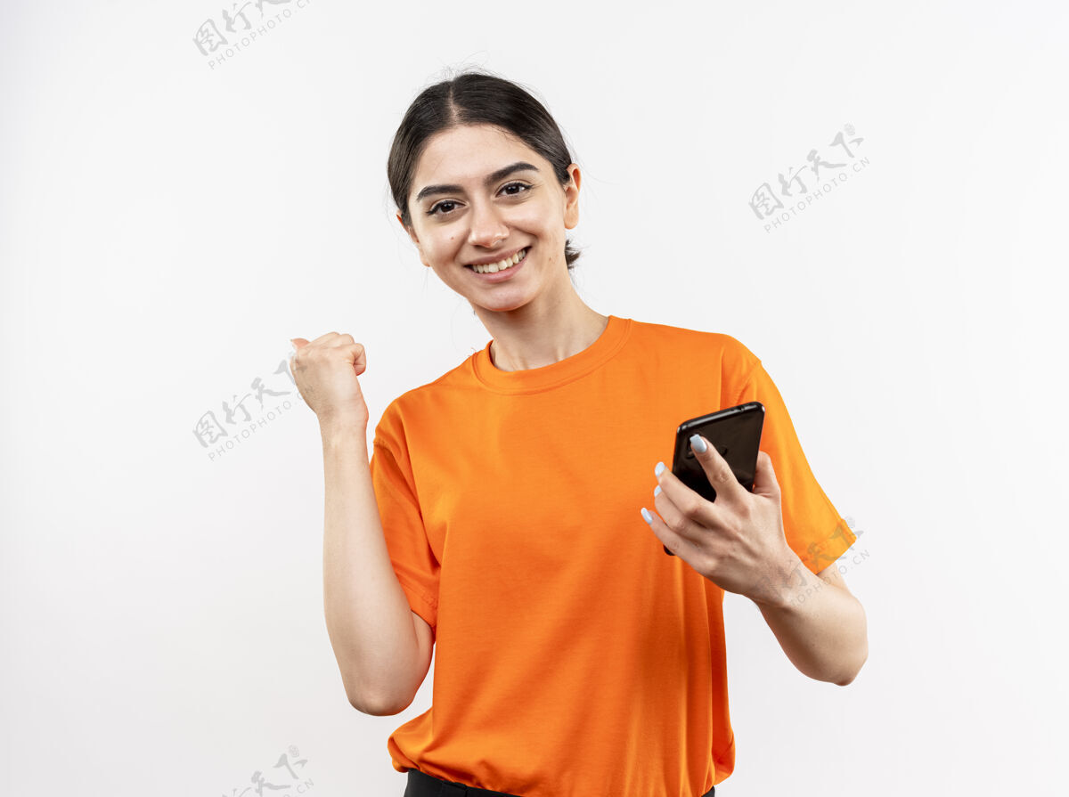 穿身穿橙色t恤的年轻女孩手持智能手机握紧拳头 高兴而兴奋地微笑着 站在白墙上为自己的成功而欢欣鼓舞微笑抱着兴奋