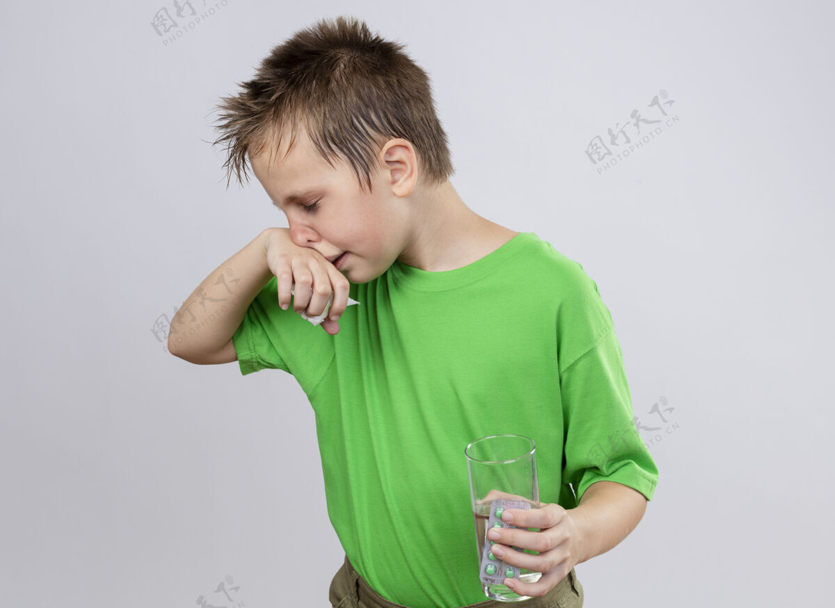 鼻子生病的小男孩穿着绿色t恤 站在白色的墙上 手里拿着一杯水和药丸 用纸巾擦鼻子 感觉不舒服站着玻璃小