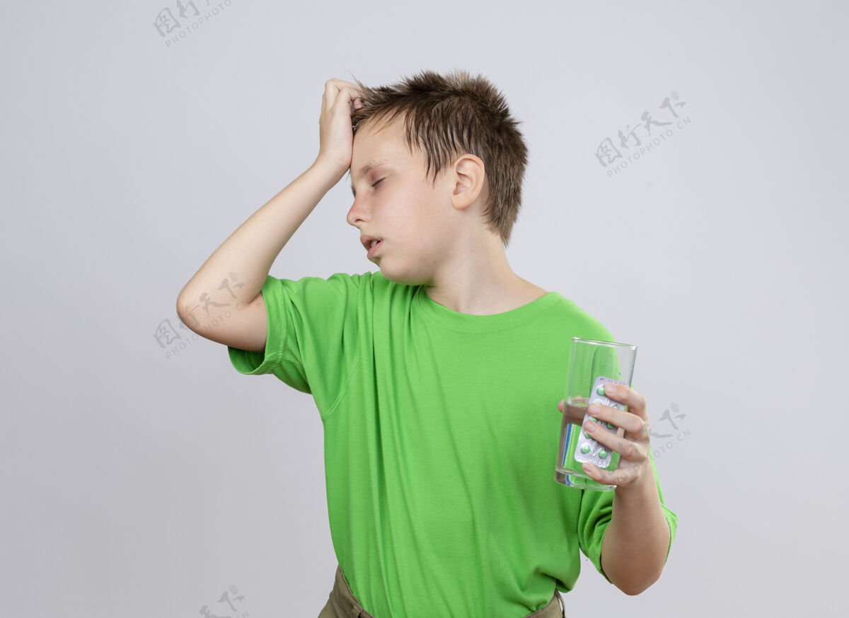 疾病生病的小男孩 穿着绿色t恤 站在白墙上 手里拿着一杯水和药片 头上挨着冻得要命抱着感冒不适