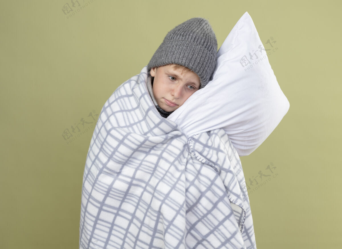 帽子生病的小男孩 戴着暖和的帽子 裹着毯子 抱着枕头 站在轻墙上感觉不舒服温暖疾病抱着