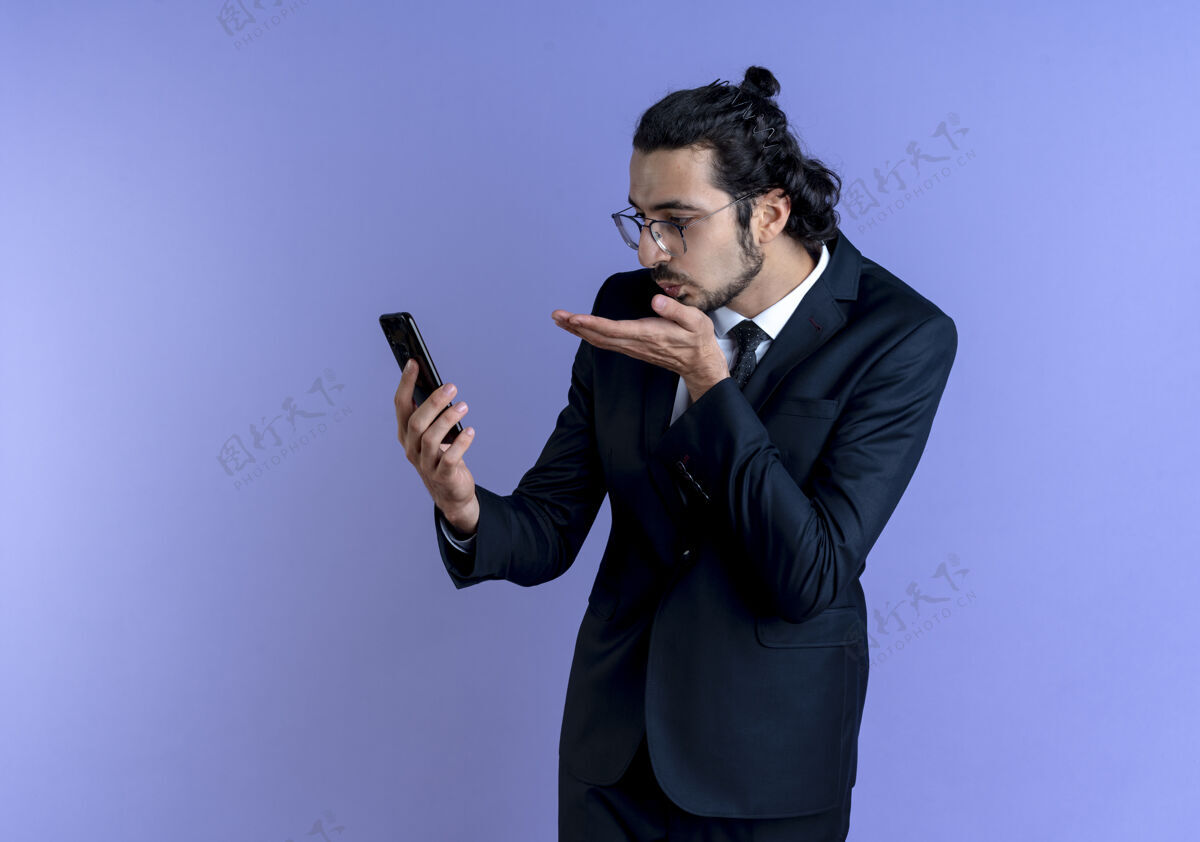 西装一个穿着黑西装戴着眼镜的商人站在蓝色的墙上看着自己的智能手机屏幕 对着电话吹着吻姿势公司亲吻
