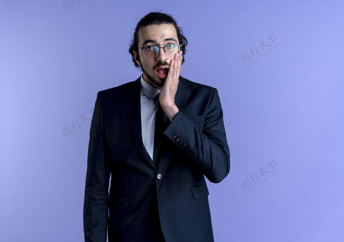 困惑一个穿着黑西装戴着眼镜的商人站在蓝色的墙上看着前面 既惊讶又困惑姿势男人工作