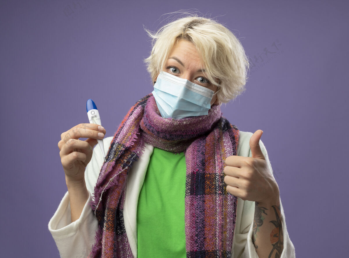 女人生病的不健康的女人 短头发 戴着暖和的围巾 戴着面罩 拿着温度计 竖起大拇指 微笑着站在紫色的墙上不健康保护短裤