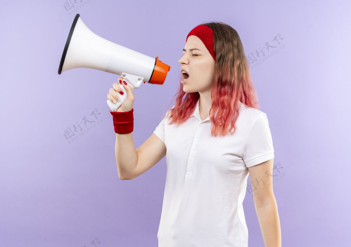 人年轻的运动型女人站在紫色的墙上对着扩音器大喊大叫 表情咄咄逼人运动装姿势喊叫