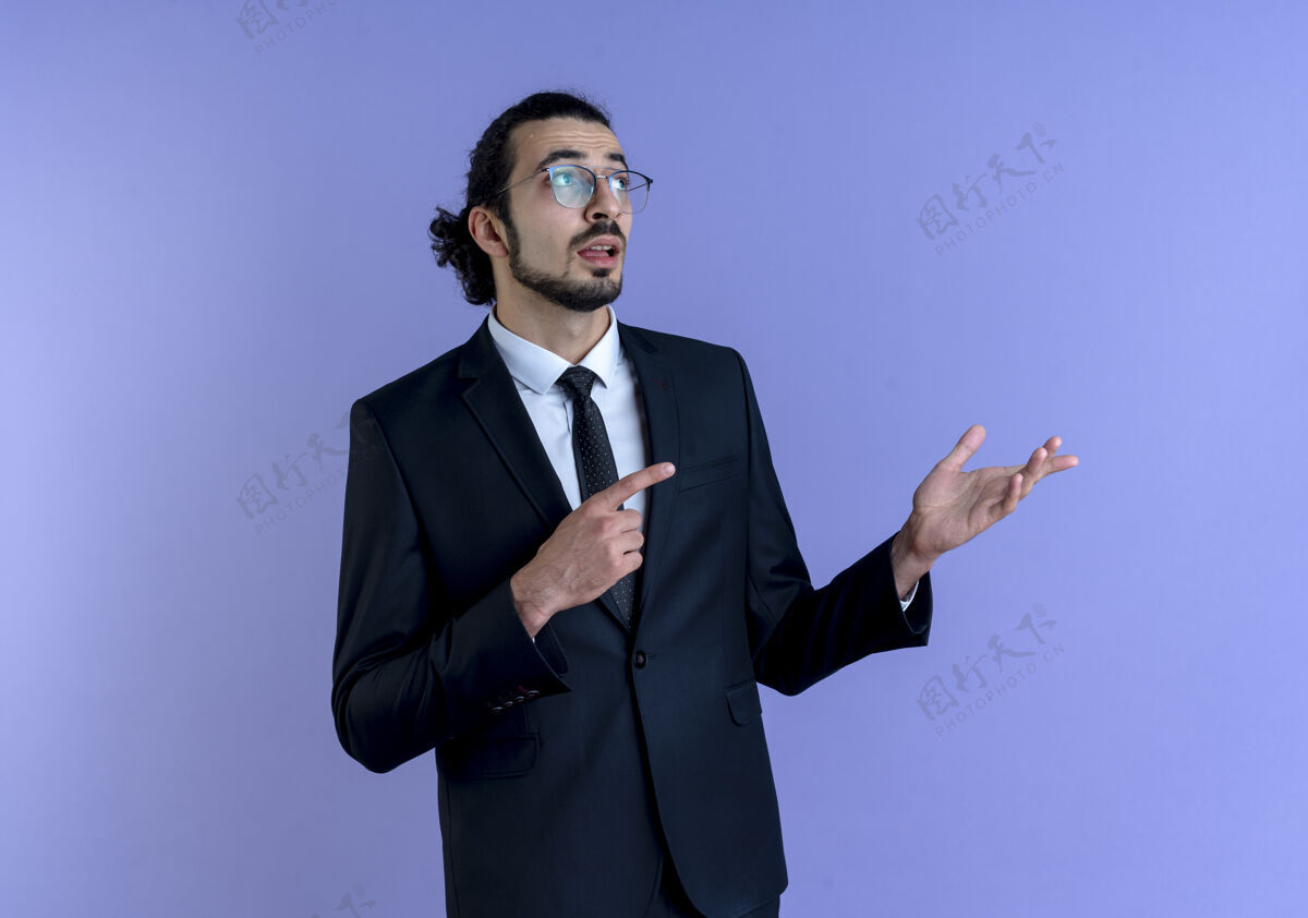 手一位身穿黑色西装 戴着眼镜的商人一边看一边用食指指着一边 他的手臂站在蓝色的墙上看指向工作
